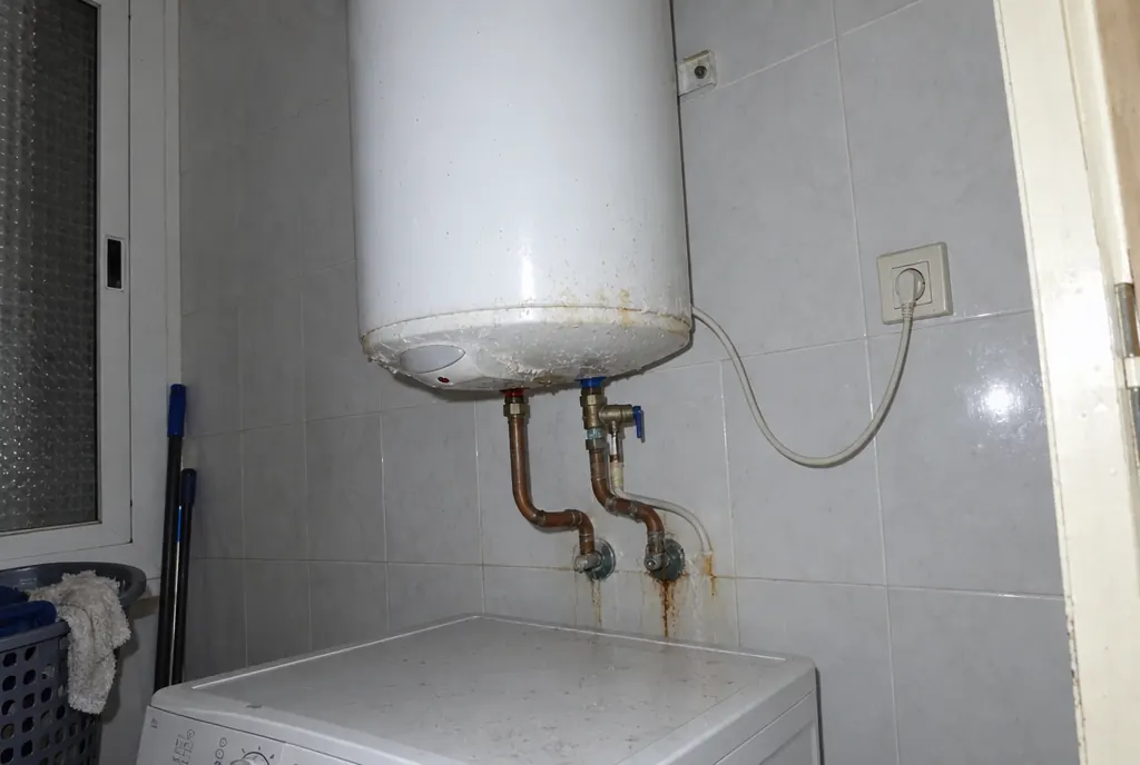 Termo eléctrico colgado sobre una lavadora, con tuberías de cobre, válvula y manchas de óxido debajo
